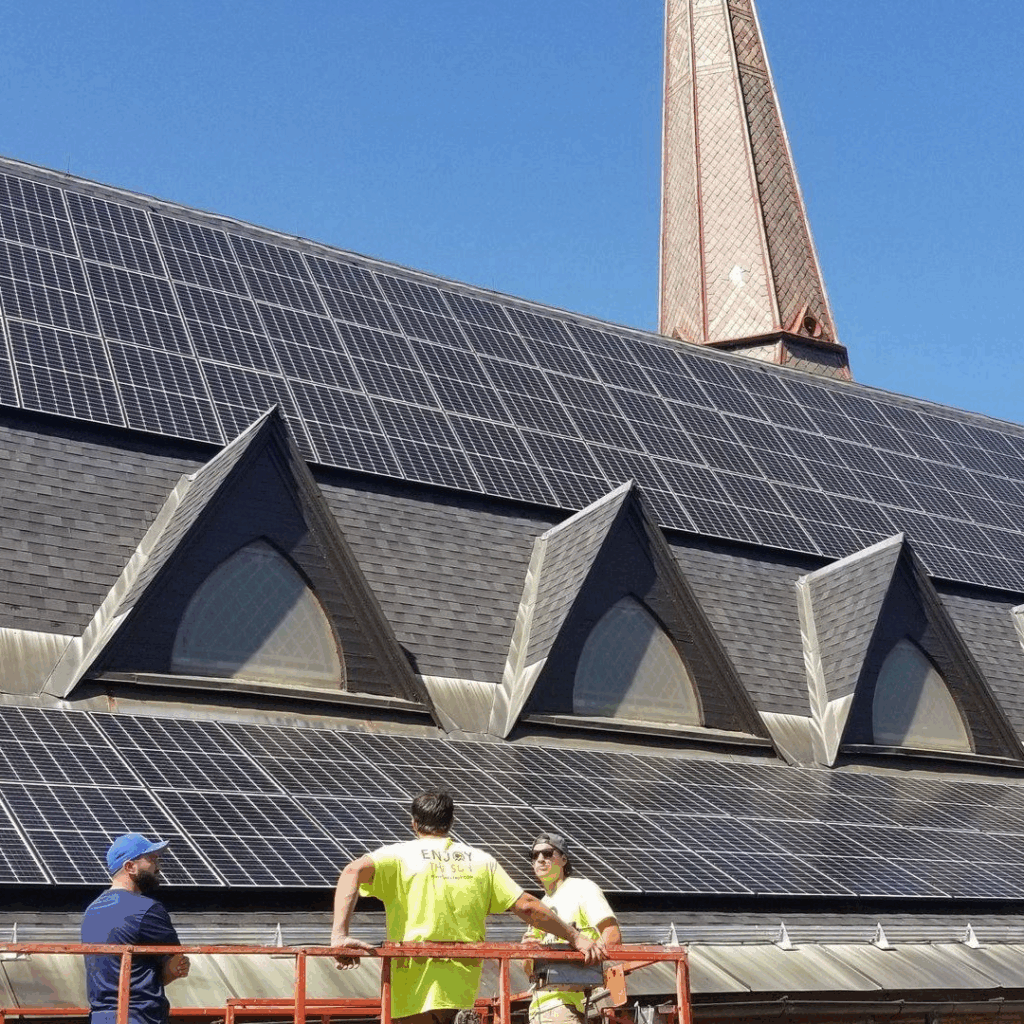 Three workers stand in front of the roof of 34 Court Stree, Keene NH, covered with solar panels, with a tall spire visible in the background under a clear blue sky.