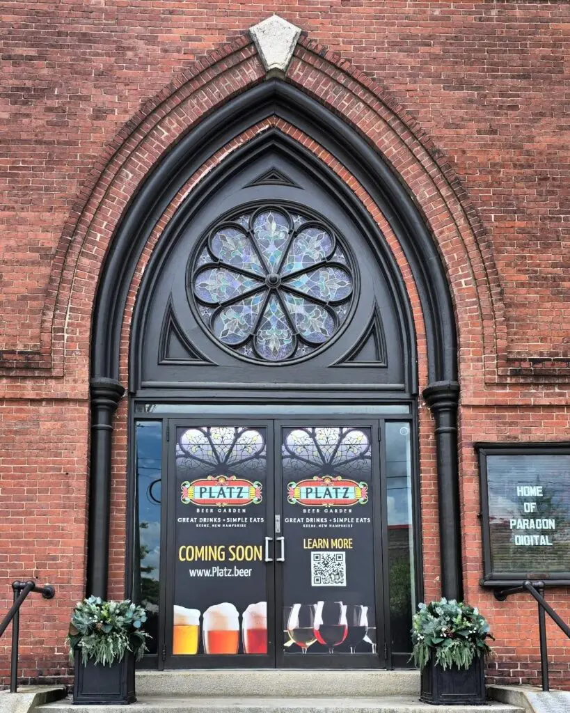 Front doors of 34 Court Street, Keene NH, marked "Platz Beer Garden", advertising a coming soon business with images of drinks, stained glass window above, and potted plants on either side of the entrance.