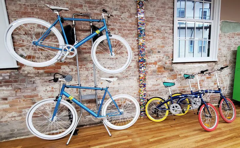 Four bicycles, two on a rack and two on the floor, against a brick wall with a colorful vertical art piece; a large window provides natural light.