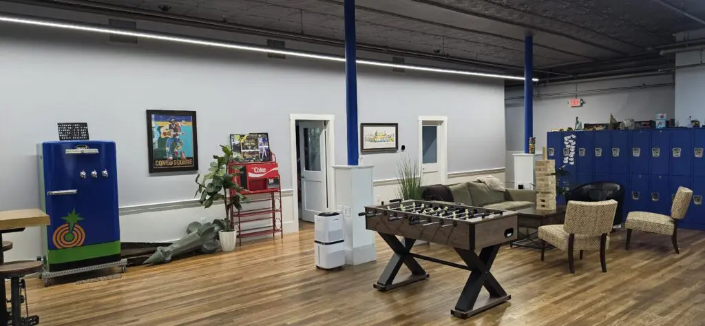 A recreational space with a retro kegerator, foosball table, and seating area with Jenga set up, against blue lockers.